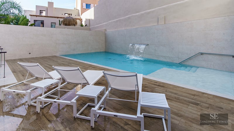 Turquoise pool with integrated waterfall, white lounge chairs on wooden decking, and privacy stone walls.