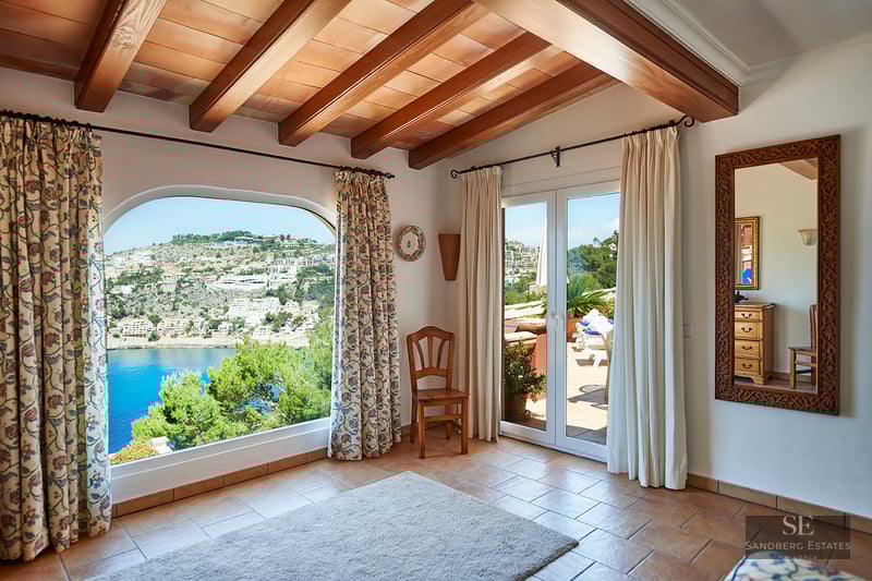 Interior room with exposed wood beams, terracotta floors, and a large arched window overlooking a coastal bay.