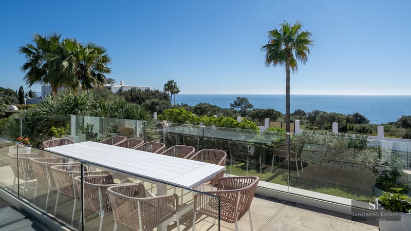 A spacious outdoor terrace with a large white table, stylish chairs, and a glass railing overlooking the sea and palm trees.