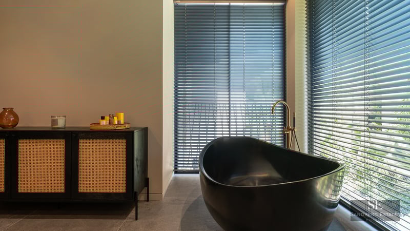 Luxury bathroom featuring a black oval bathtub, gold faucet, and a black rattan sideboard next to windows with blinds.