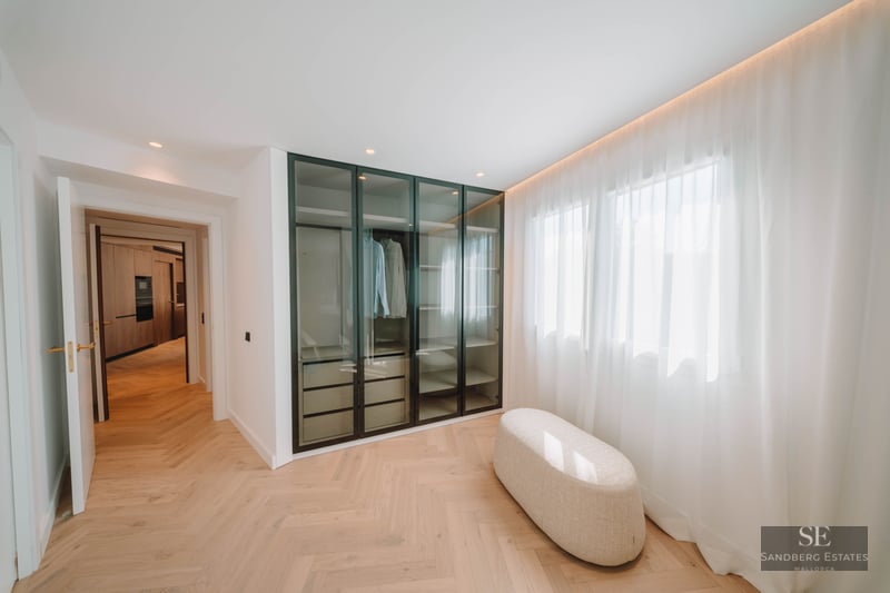 Modern walk-in closet with herringbone wood floors, glass wardrobe, and a central ottoman next to a window.