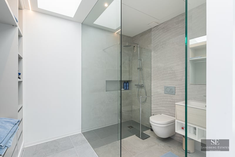 Minimalist bathroom featuring grey stone tiles, a glass walk-in shower, wall-hung toilet, and a bright skylight.