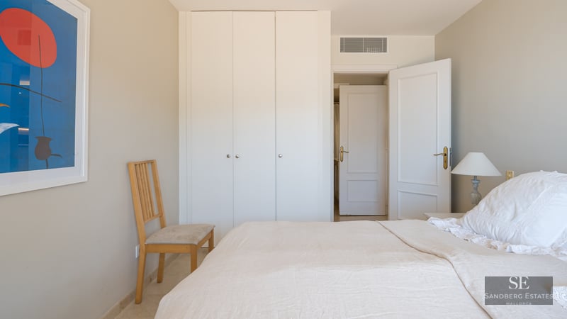 Bright bedroom featuring a white bed, large white built-in closets, a wooden chair, and abstract art on the wall.