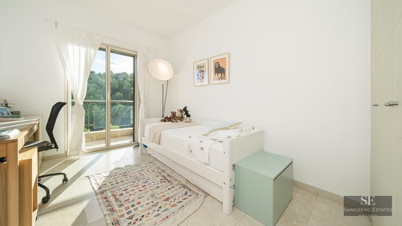 White bedroom featuring a single bed, desk, sliding glass door to a balcony, and a geometric rug.