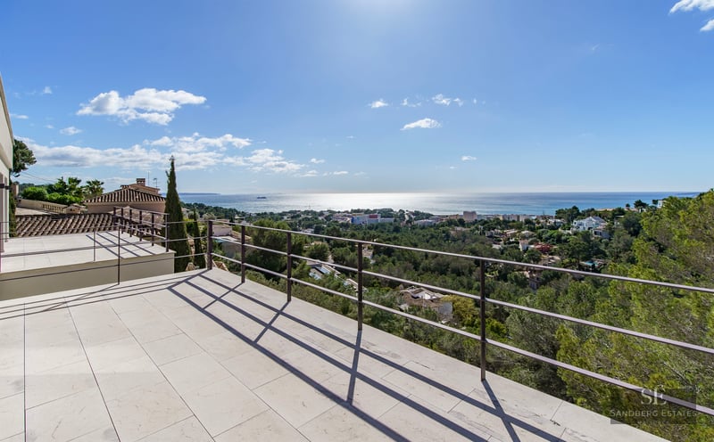 Large white stone terrace with a black metal railing overlooking a panoramic view of the Mediterranean Sea and pine trees.