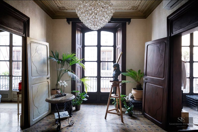 Grand entrance with tall wooden doors, mosaic floors, a crystal chandelier, and indoor plants by a balcony window.