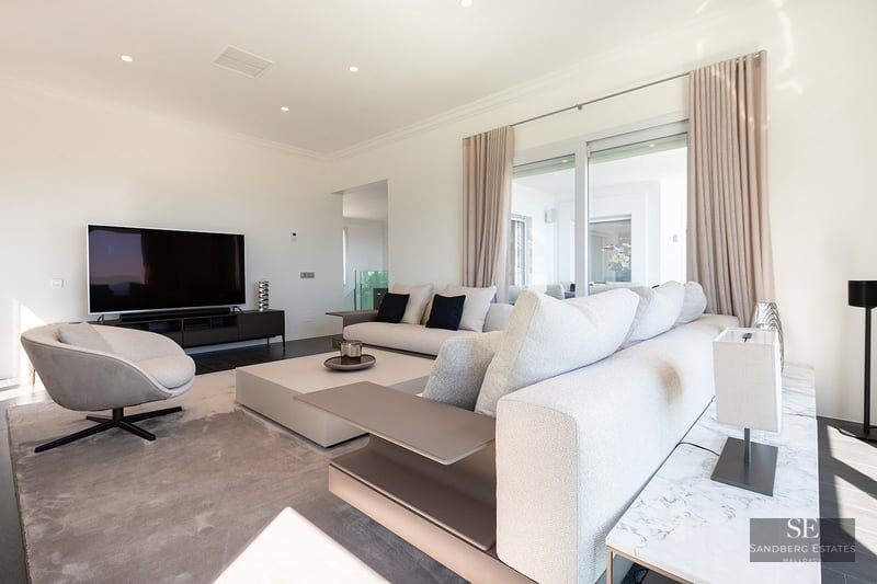 Contemporary living room featuring a large beige sofa, a designer armchair, and a flat-screen TV in a bright space.