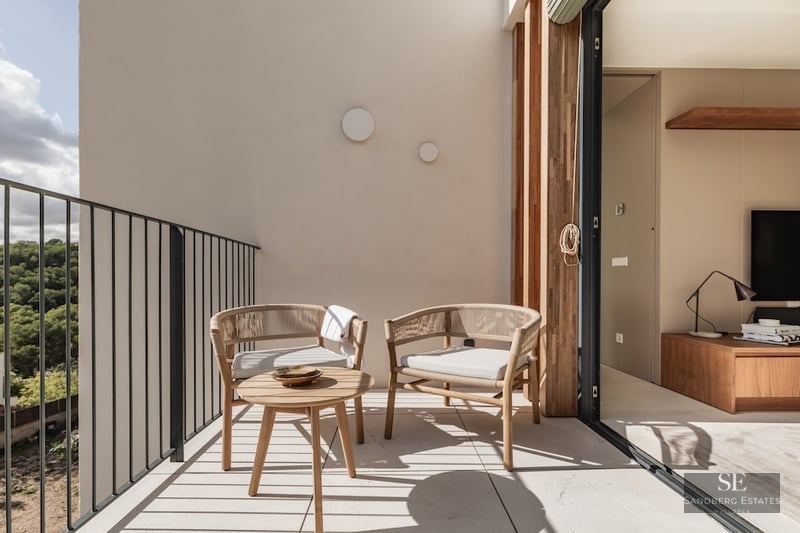 Sunlit balcony with designer woven chairs and a wooden coffee table overlooking a lush green forest.