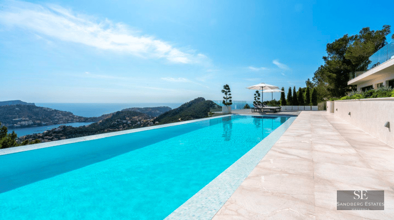 Turquoise infinity pool overlooking the sea and mountains, surrounded by light stone tiles and white umbrellas.