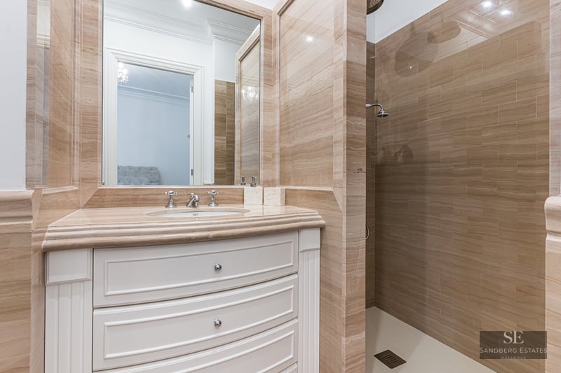 Luxury bathroom featuring beige travertine tile walls, a white curved vanity with marble top, and a walk-in shower.