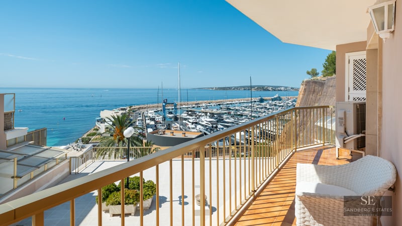 Balcony with a white wicker chair and gold railing overlooking a marina full of boats and the blue Mediterranean Sea.