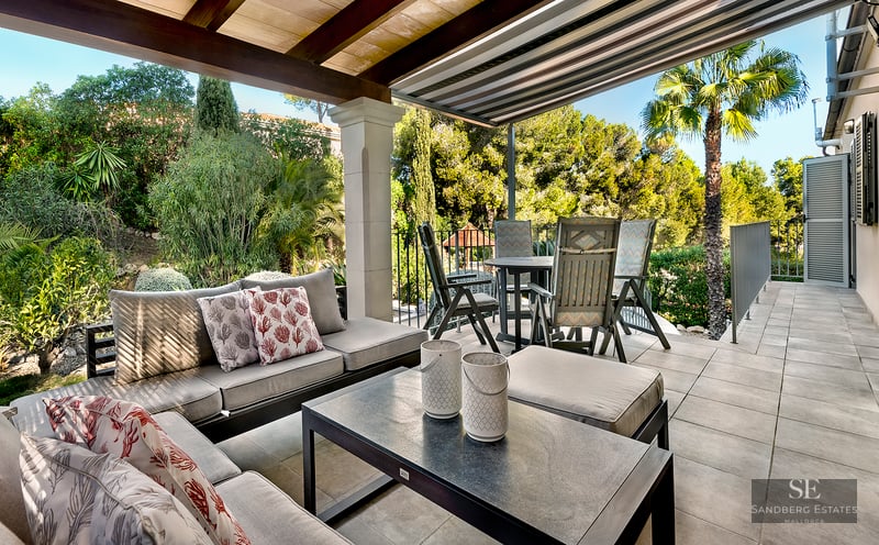 Covered outdoor terrace with lounge sofas, dining table, and view of a lush Mediterranean garden under a bright sky.