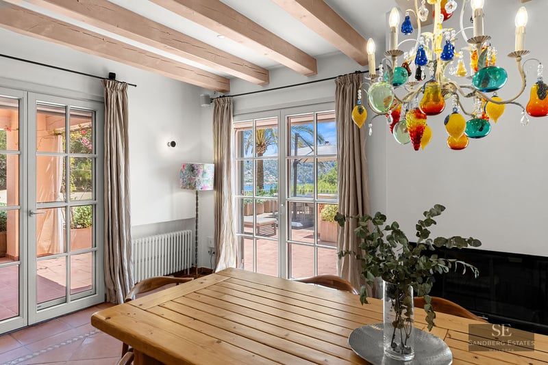 Interior dining room featuring a wooden table, colorful glass chandelier, exposed beams, and doors to a sunny terrace.