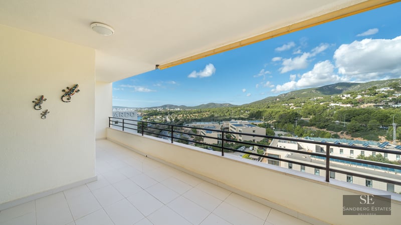 Spacious white tiled balcony with a black metal railing overlooking green hills and a blue sky with white clouds.