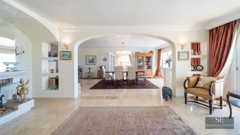 Spacious living area with marble floors and an archway framing a formal dining room with a chandelier and classic rugs.
