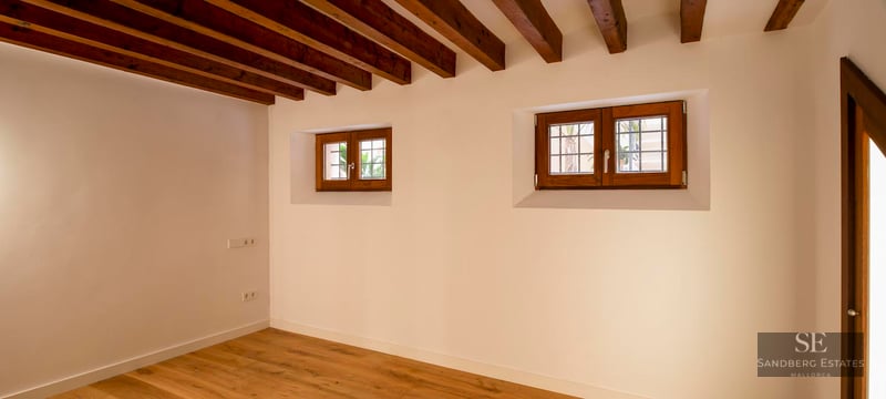 Empty room featuring dark wood-beamed ceiling, white walls, and small wooden windows with hardwood floors.