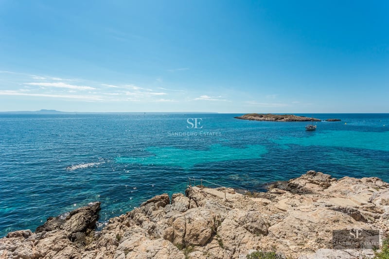 Panoramic view of turquoise Mediterranean waters from a rocky shore with a small island and boat in the distance.