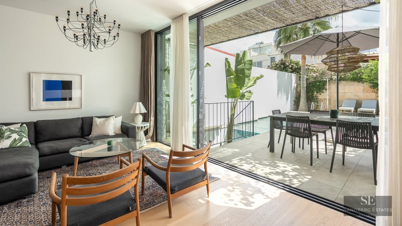 Bright living room with dark sofa and wooden chairs opening to a sunny terrace with a swimming pool and dining area.