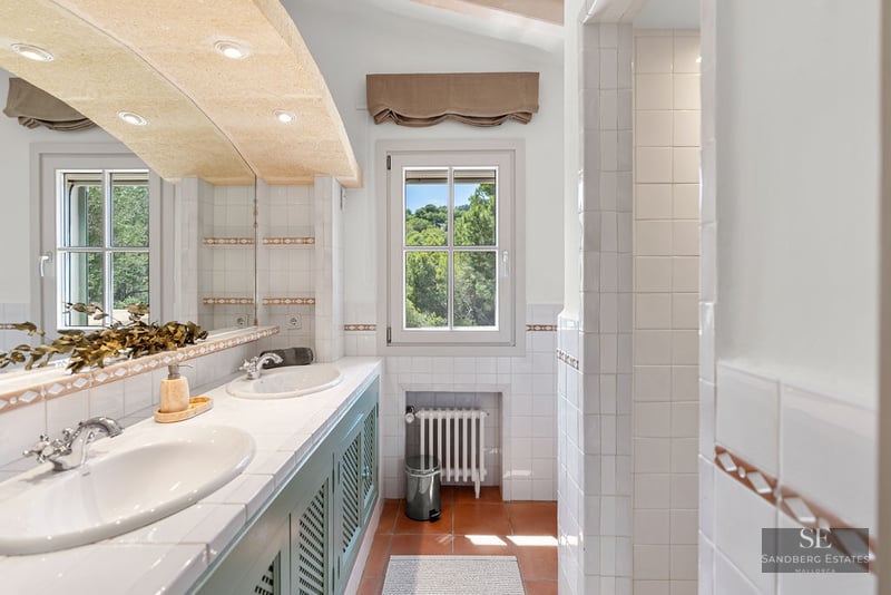 Bright bathroom featuring double white sinks, green cabinetry, terracotta tile floors, and a window viewing lush trees.