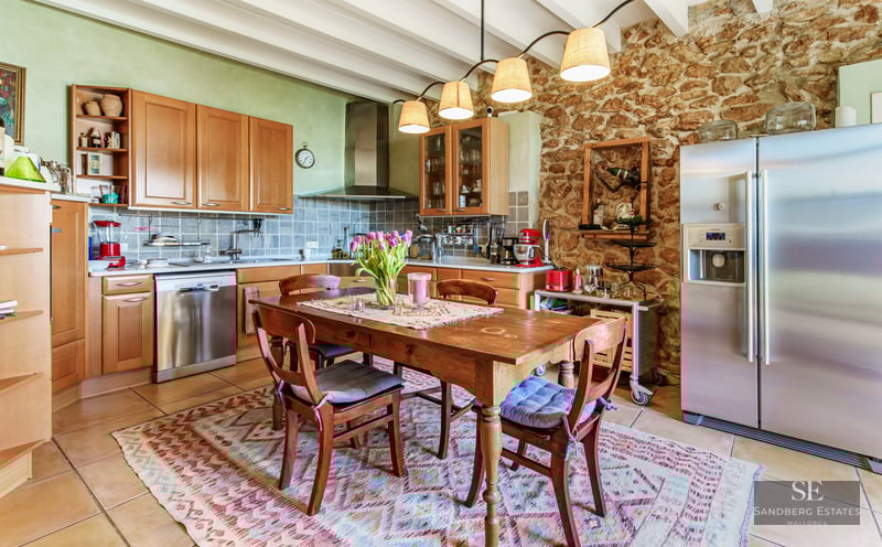 A rustic kitchen featuring an exposed stone wall, wooden cabinets, a central dining table, and wooden ceiling beams.