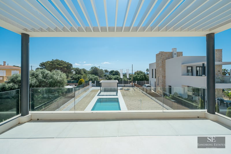 View from a modern balcony with a white pergola and glass railing overlooking a rectangular swimming pool and garden.