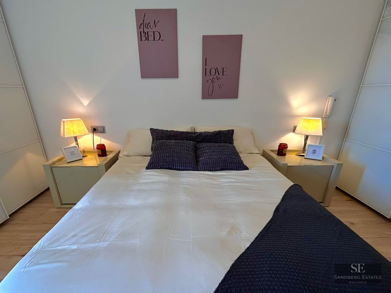 Neatly made bed with white linens and purple pillows between two glowing bedside lamps in a modern room.