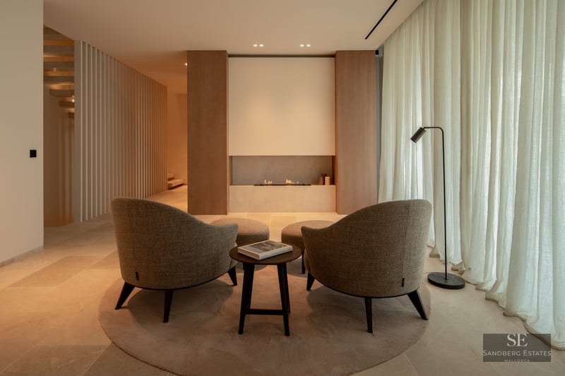 Minimalist lounge featuring two beige armchairs facing a built-in linear fireplace, warm ambient lighting, and wood paneling.