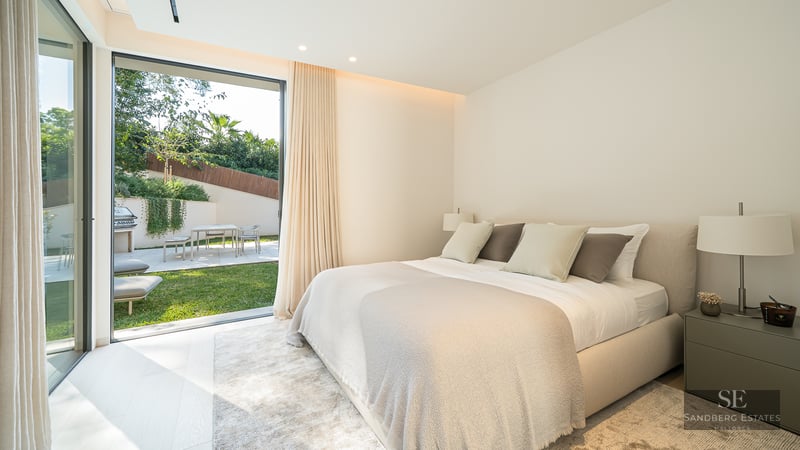A bright, modern bedroom with a large bed opening onto a sunny private garden through glass sliding doors.