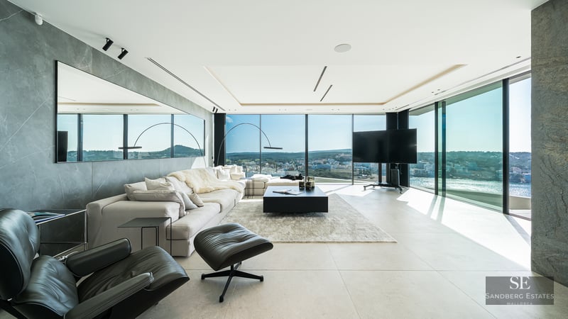 Modern living room with a large white sofa, black leather lounge chair, and floor-to-ceiling windows showing a coastal view.