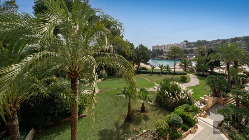 High-angle view of a tropical garden with palm trees, green lawn, and stone paths overlooking a blue sea cove.
