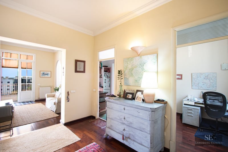 Central hallway with dark wood floors connecting a living room, kitchen, and office in a bright apartment.
