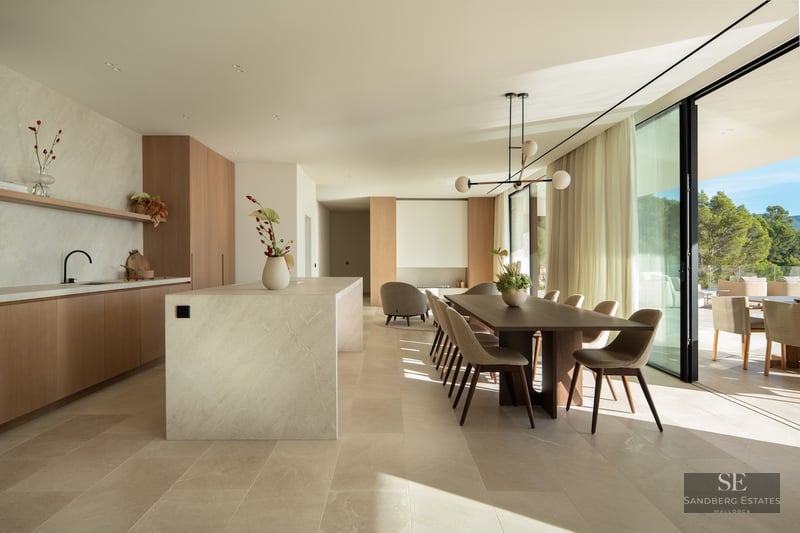 Modern dining room with a wooden table, stone kitchen island, and large floor-to-ceiling windows overlooking a forest.