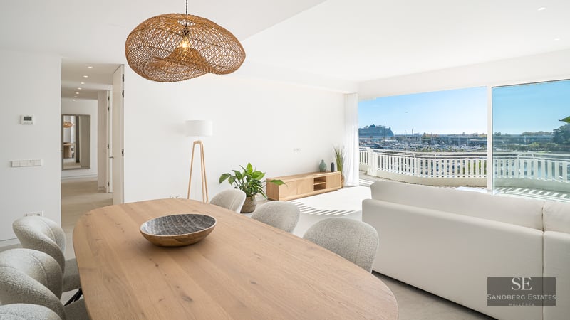 Bright living and dining room with a large wooden table, white sofa, and a balcony overlooking a marina.