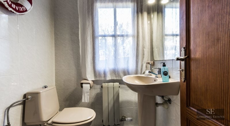 Small bathroom featuring a beige toilet and pedestal sink, a window with a sheer curtain, and light-tiled walls.