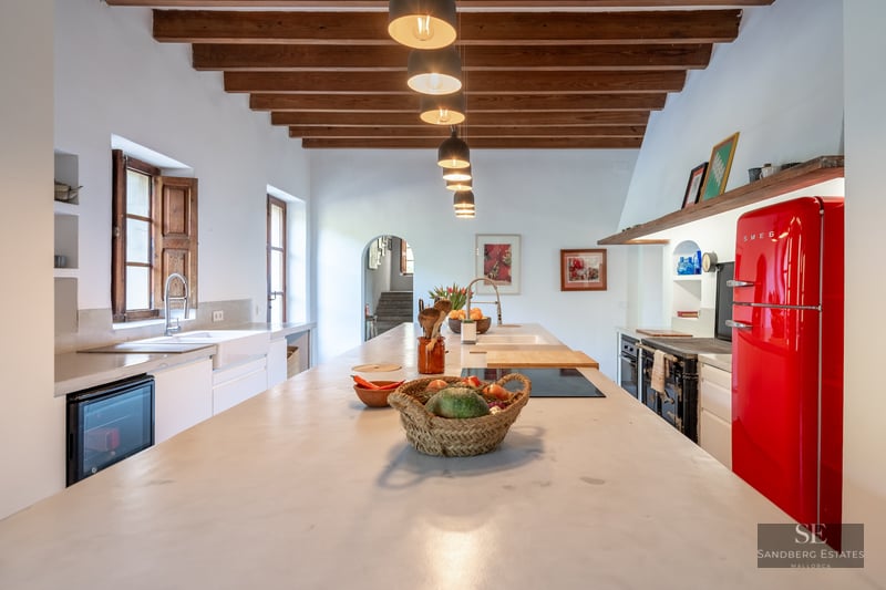 Bright kitchen with wood-beamed ceiling, long stone island, and a vibrant red retro refrigerator.