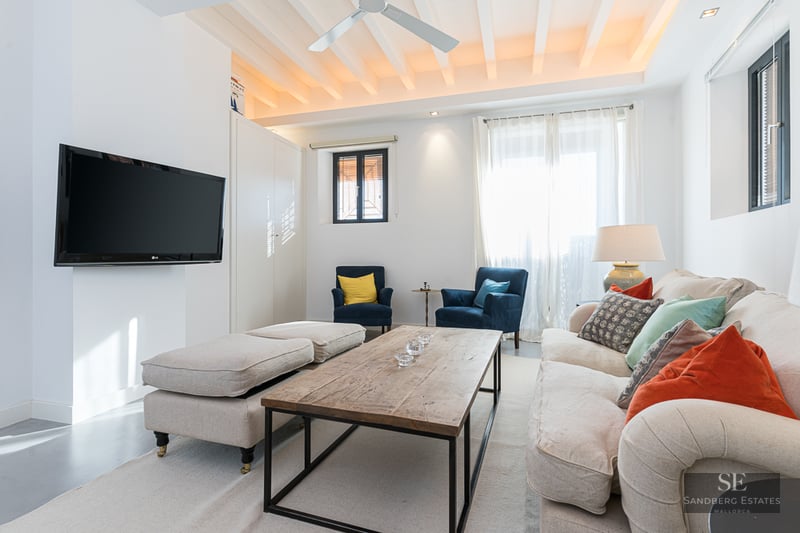 Bright living room with white beamed ceiling, beige sofa with colorful cushions, and wall-mounted flat-screen TV.