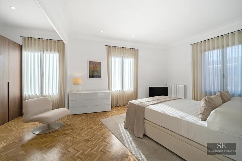 Bright bedroom with white walls, herringbone parquet, a double bed, designer chair, and sheer curtains.