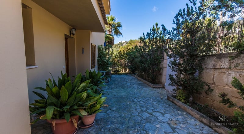 Stone-paved garden pathway alongside a beige Mediterranean house with lush green plants and terracotta pots.