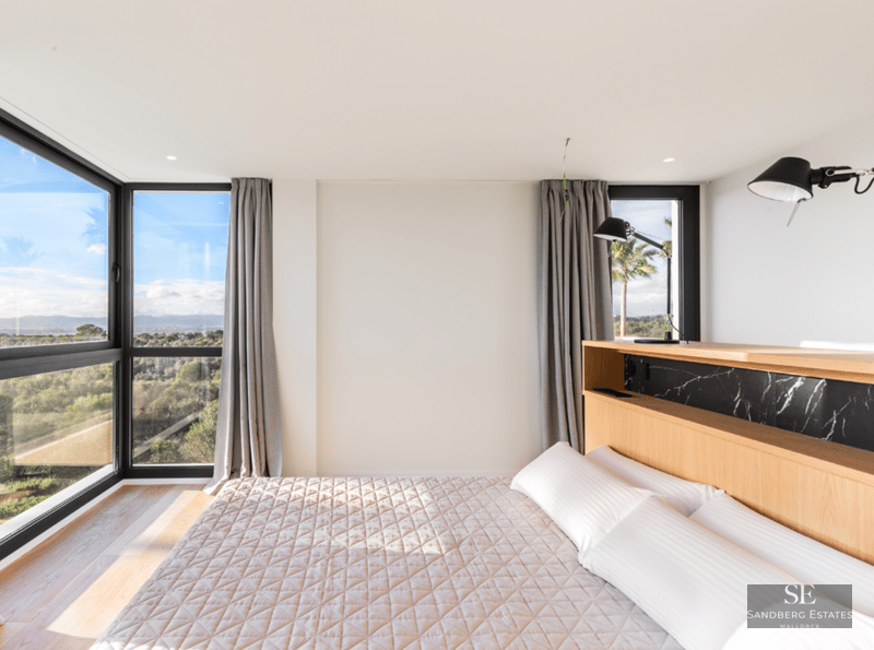 Bright bedroom with large windows overlooking trees, wooden headboard with black marble, and white bedding.