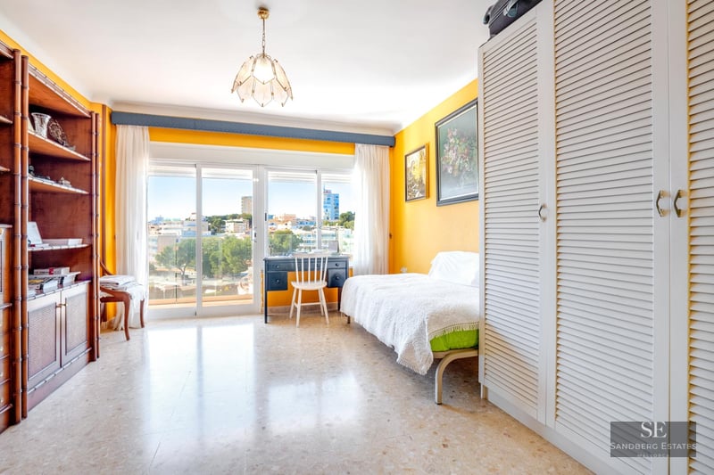 A sunny bedroom featuring yellow walls, a white slatted wardrobe, a desk by a large window, and city views.
