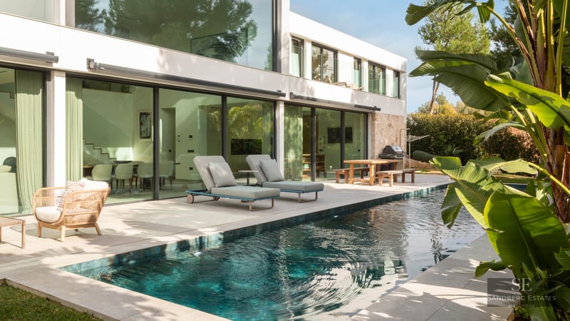Piscine turquoise à côté d'une villa blanche moderne avec de grandes baies vitrées et des transats.