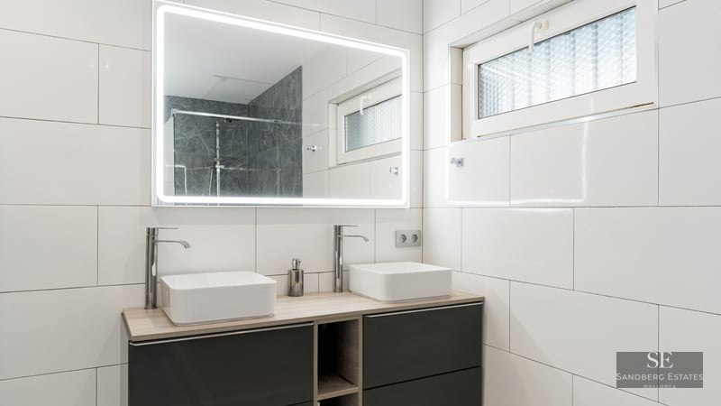 Salle de bain moderne avec deux vasques carrées, un grand miroir éclairé et des murs en carrelage blanc.