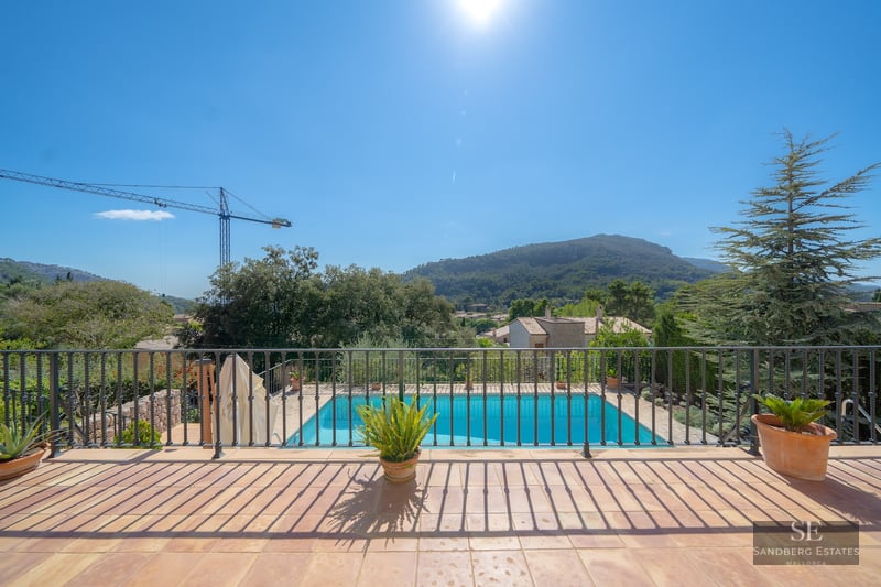 View from a terrace with iron railing overlooking a blue swimming pool and green mountains under a bright sun.