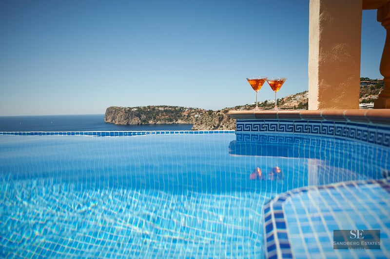Close-up of a blue tiled infinity pool with two orange cocktails on the ledge overlooking the sea and cliffs.