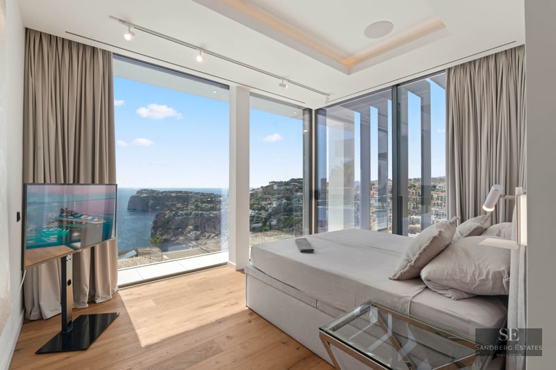 Modern bedroom with floor-to-ceiling windows showing a panoramic view of the sea and coastline.