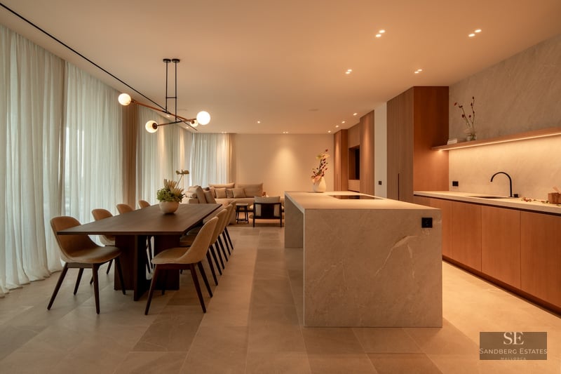 Warmly lit open-plan room with a stone kitchen island, dining table, and sofa behind soft curtains.