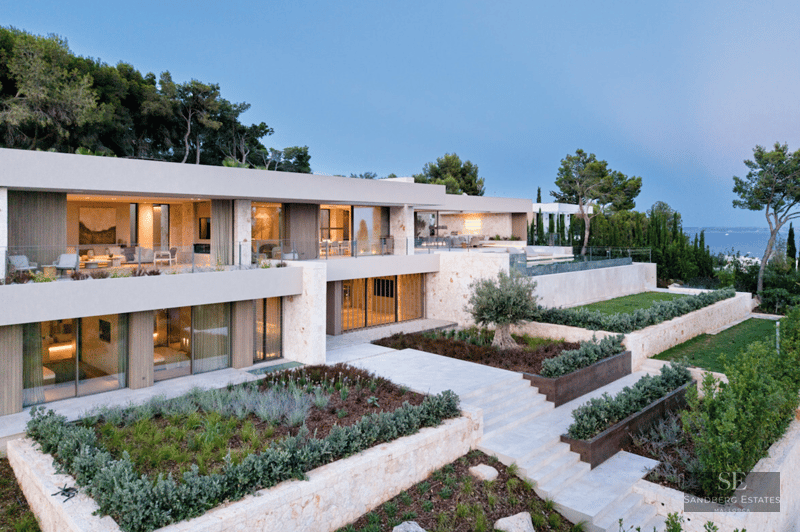 Modern multi-level villa with stone walls, large windows, and tiered Mediterranean gardens overlooking the sea at dusk.
