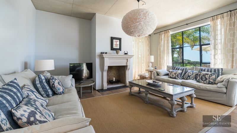 Spacious living room with beige sofas, stone fireplace, and large windows viewing garden palms.