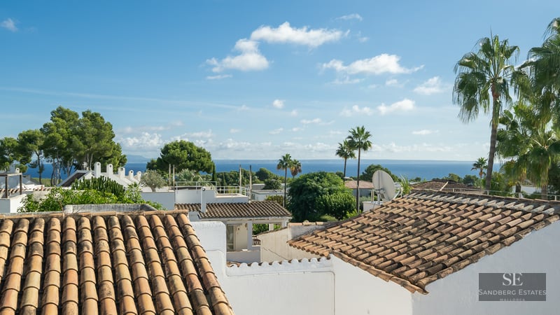 Elevated view of terracotta roofs, white walls, and palm trees with the blue sea in the background under a sunny sky.