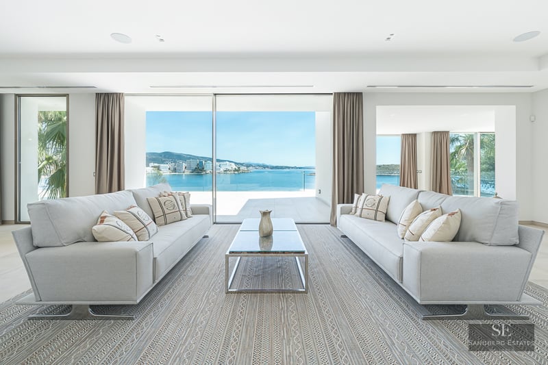 Bright living room with two grey sofas, a glass coffee table, and floor-to-ceiling windows overlooking the ocean.